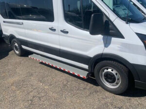 A white passenger van is parked on asphalt, with a black side mirror and a striped step rail along the side.