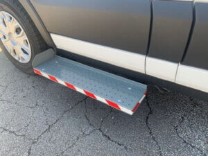 A metal running board with red and white reflectors is mounted below the door of a parked vehicle on a cracked asphalt surface.