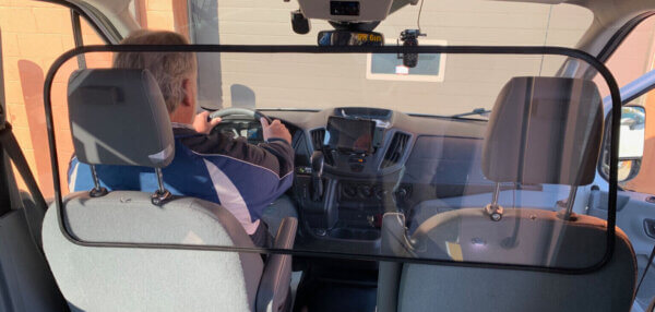 A driver sits in a vehicle separated from the back seats by a clear plastic partition attached to the front seat headrests.