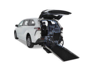 A white Toyota Sienna Hybrid minivan with its rear hatch open, showing a wheelchair on a rear-entry ramp leading into the vehicle’s interior.