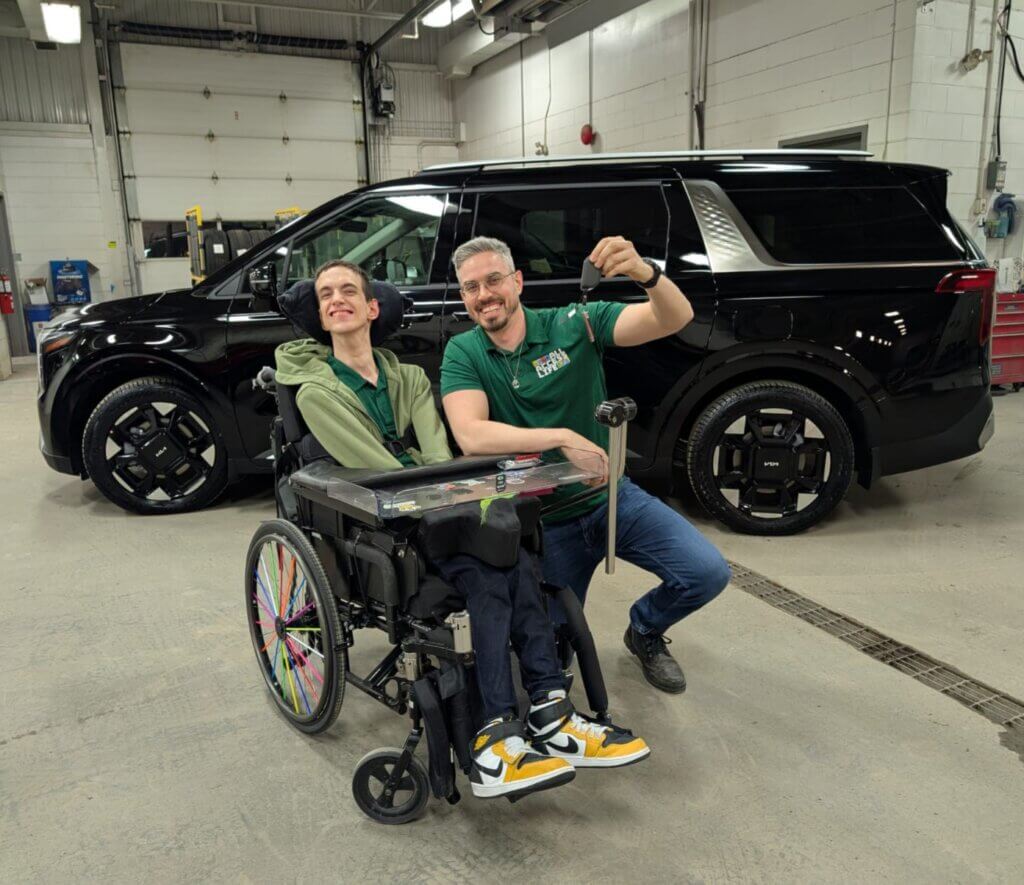 Two men, one in a wheelchair and the other kneeling beside him holding car keys, pose in front of a black SUV inside a garage, celebrating the Driverge and All Access Life partnership.