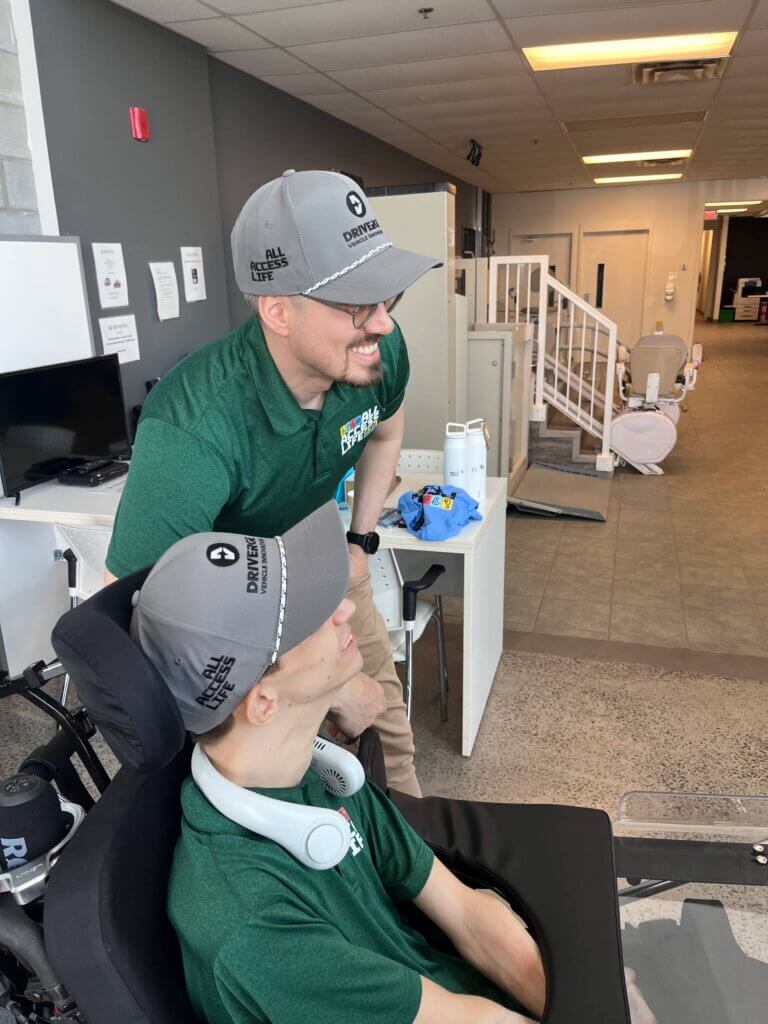Two people wearing matching green shirts and gray hats, representing All Access Life, are inside an office space. One is seated in a wheelchair while the other stands beside him, highlighting partnership and inclusion as they both face the same direction.