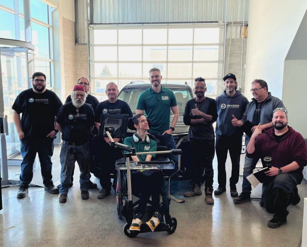 A group of ten people, including one person in a wheelchair, pose together and smile in a garage with a car and large windows in the background, celebrating the Driverge partnership with All Access Life.