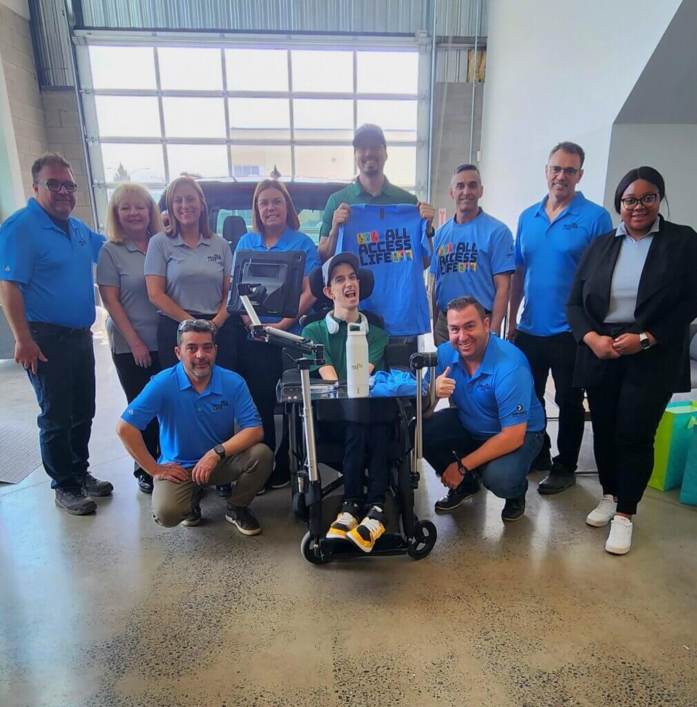 A group of people, some in blue shirts, pose indoors with a smiling child in a wheelchair holding a shirt that reads “ALL ACCESS LIFE,” celebrating the partnership between All Access Life and Driverge.