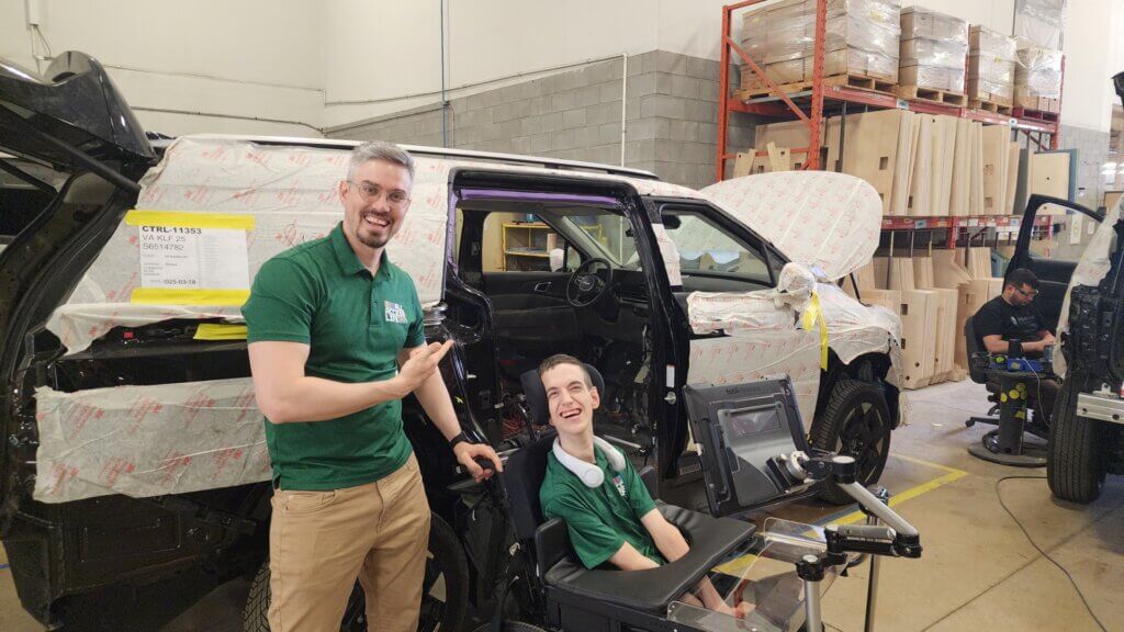 Two people smile next to a partially assembled vehicle in a workshop, showcasing innovative assistive technology. This scene celebrates the partnership between All Access Life and Driverge, with another person working in the background.