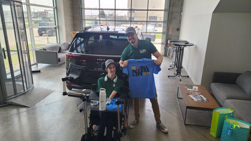 Two people in green shirts pose indoors by a black Kia SUV; one is in a wheelchair, and the other holds a blue “All Access Life” T-shirt, celebrating the partnership between All Access Life and Driverge.