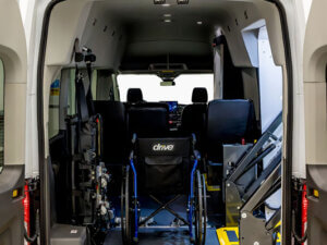 The interior of a wheelchair-accessible van is shown from the back, with a secured wheelchair, ramp, and seating visible.