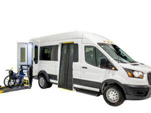 A white passenger van with a side door open and a wheelchair lift extended, loading a blue wheelchair into the vehicle.